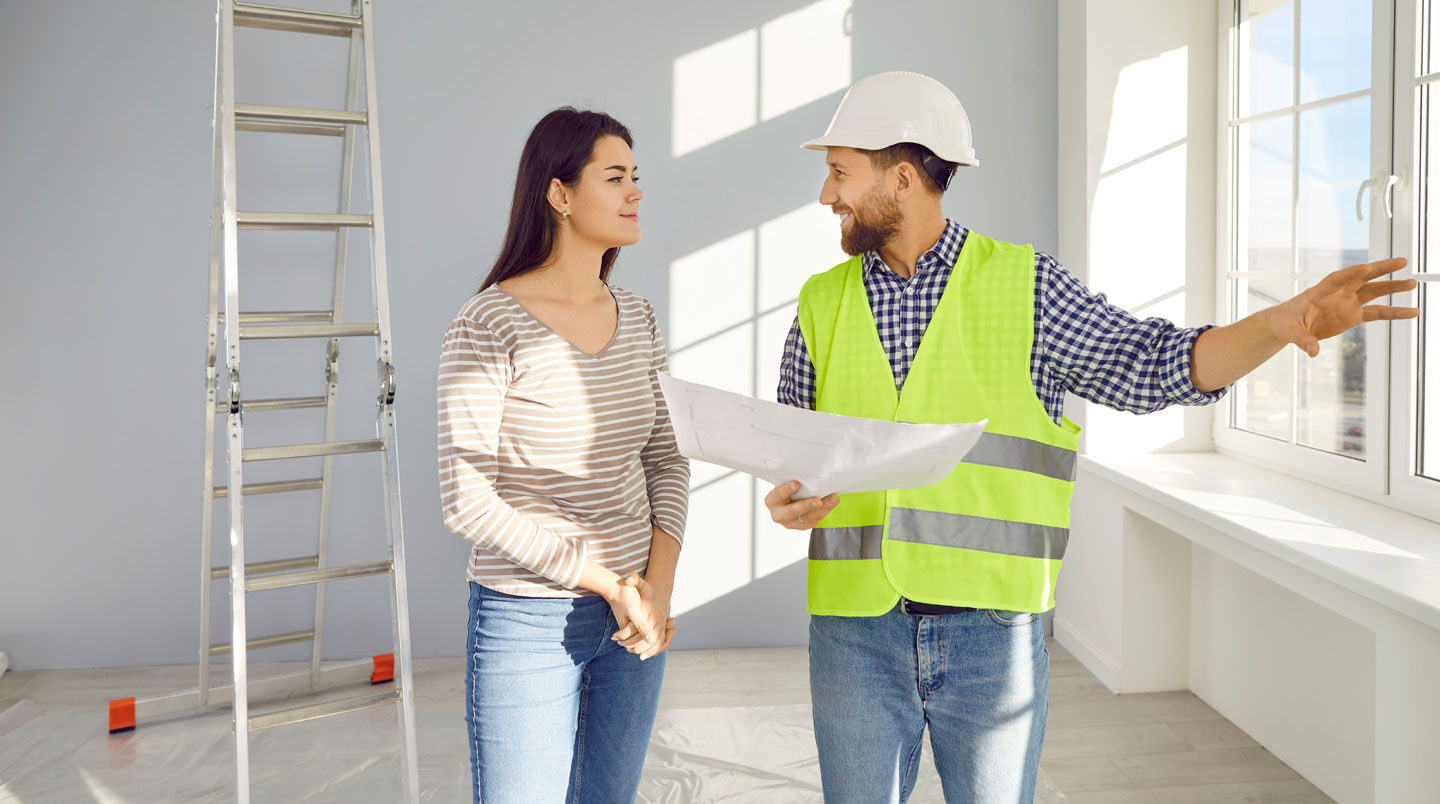 Réunion de chantier entre couple de clients et professionnel de la construction autour de plans de maison, site de construction en Haute-Savoie en arrière-plan